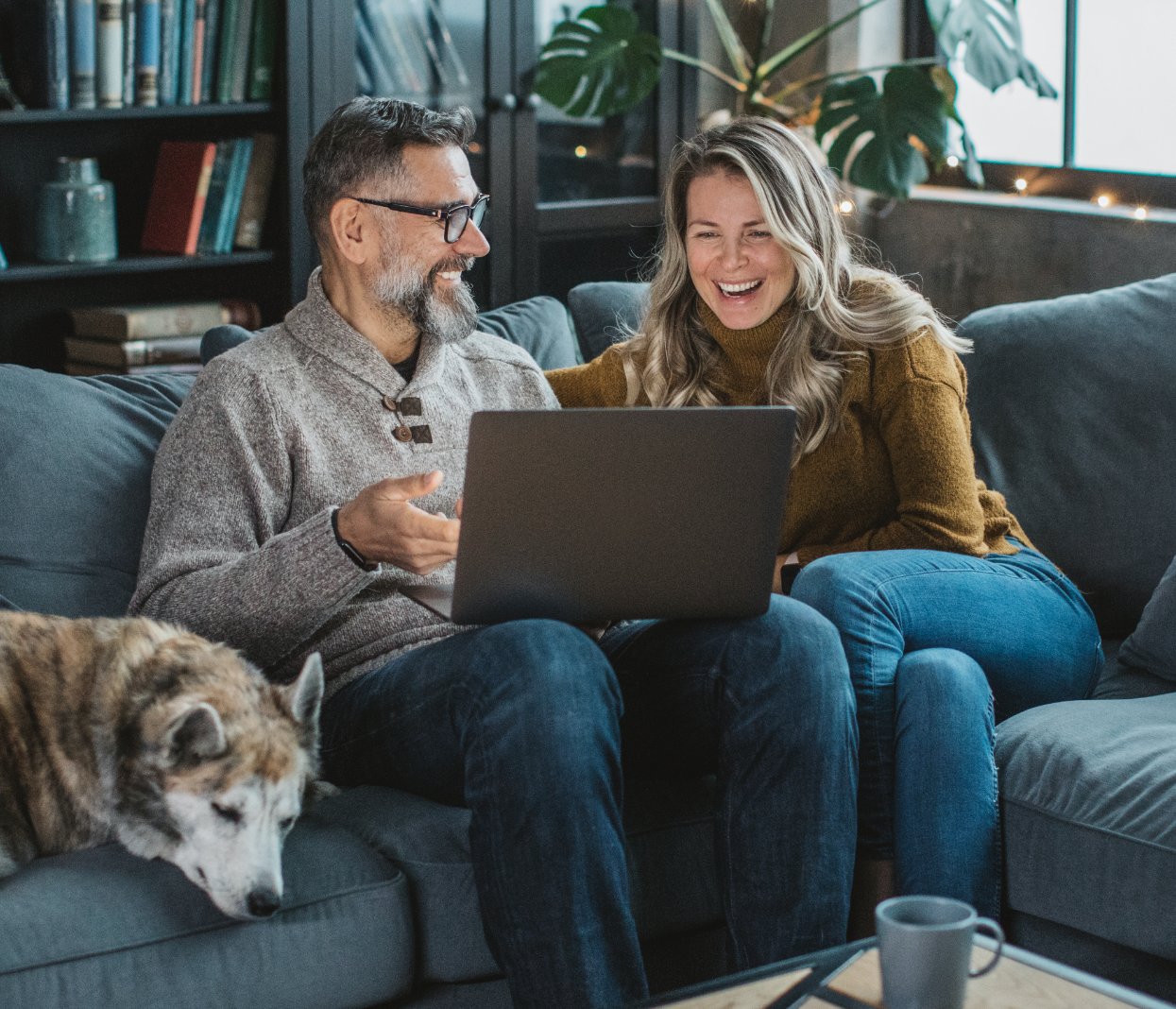 Older couple sitting on the couch with their dog, browsing the internet on their laptop