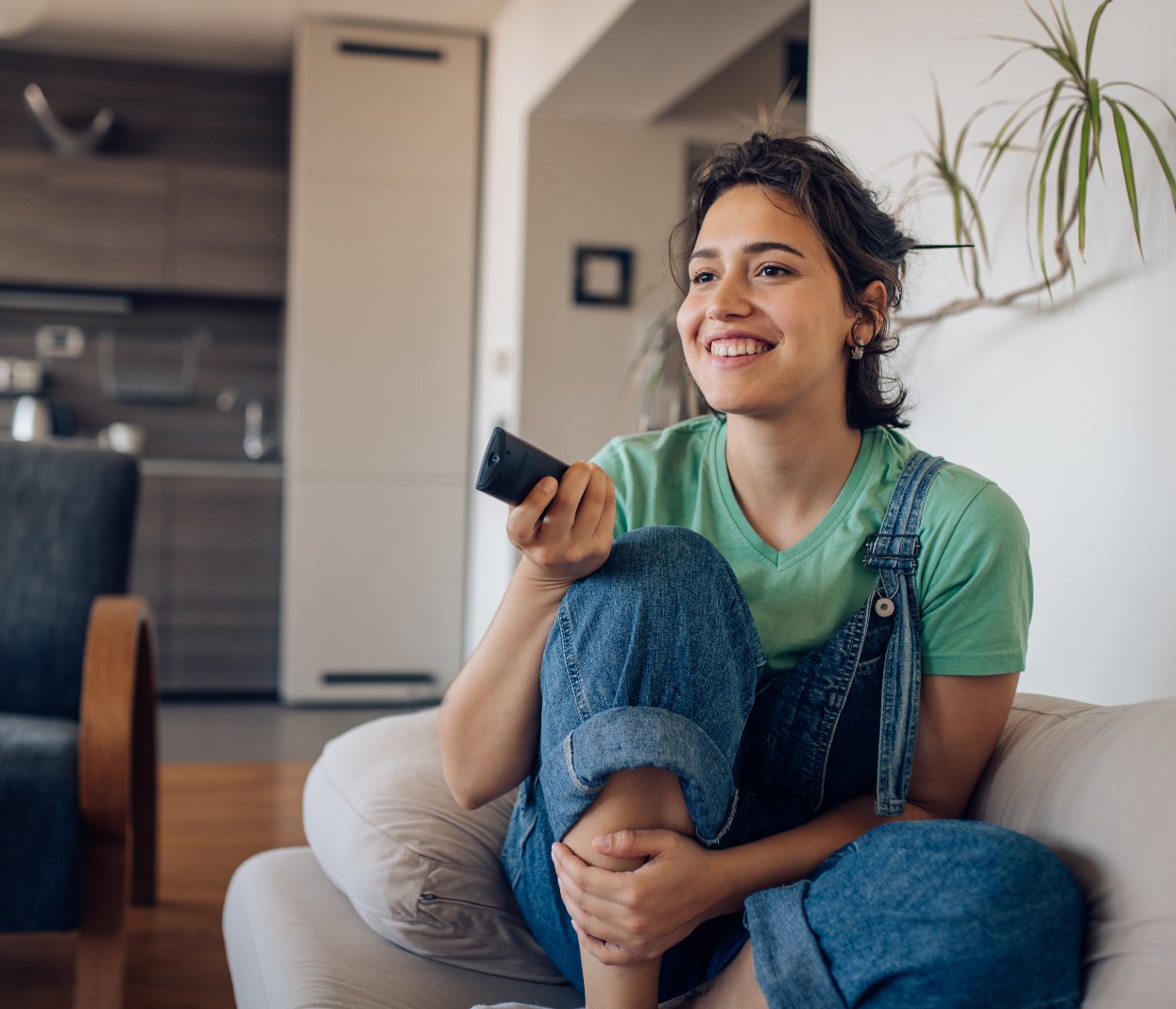 Young woman in overalls sitting on the couch watching television with a remote control in her hands