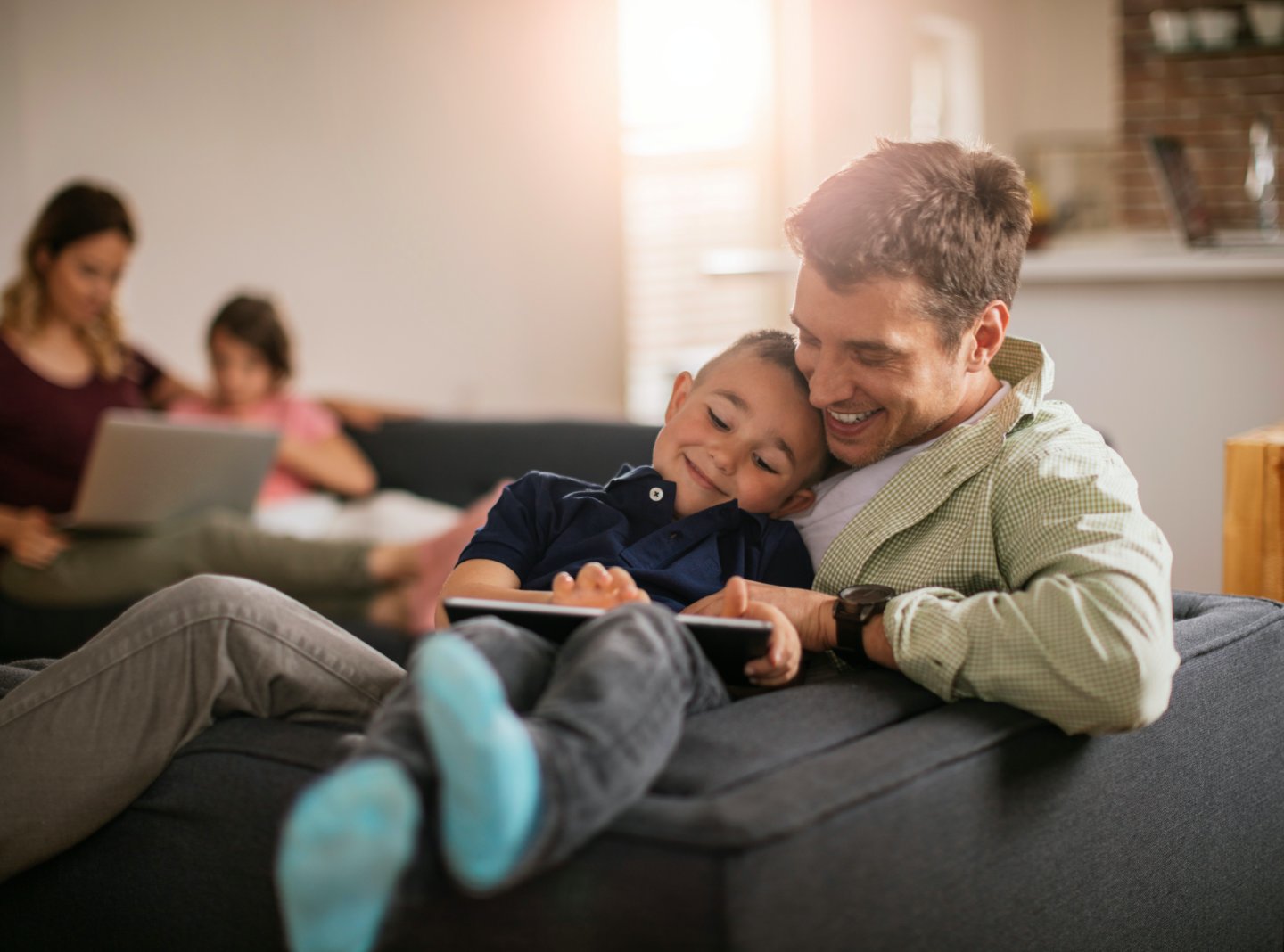 Father and son using a tablet while mother and daughter use a laptop in the background