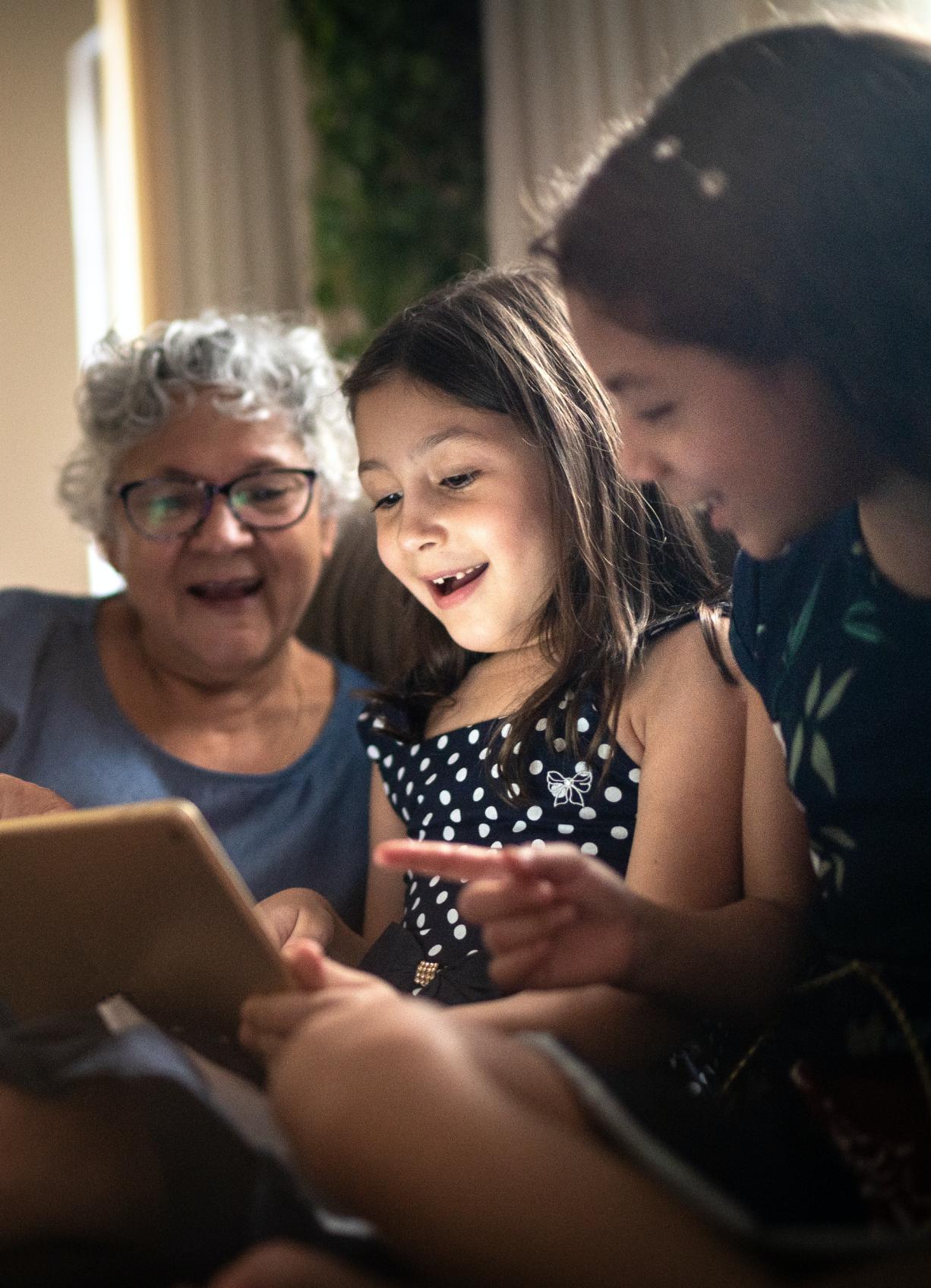 Two young girls using a tablet with grandmother watching in background